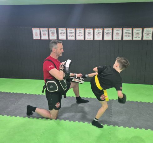 A trainer holds pads while a child performs a kick in a martial arts studio.